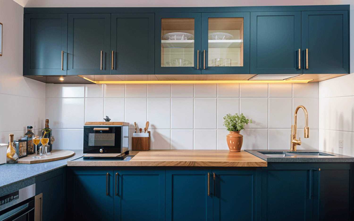 A modern kitchen with dark blue cabinets, a wooden countertop, built-in appliances, and a small plant on the counter. White tiled backsplash and gold fixtures enhance the elegant look.